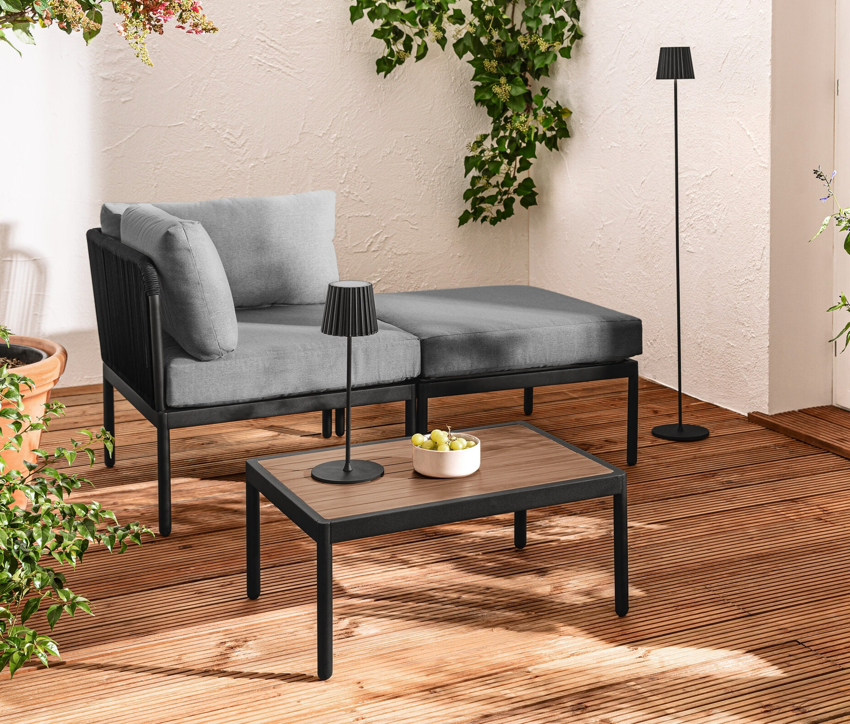 Terrasse avec lounge modulaire avec tressage textile, lampe de table et lampadaire d’extérieur à LED rechargeable noire et guirlande lumineuse à LED.