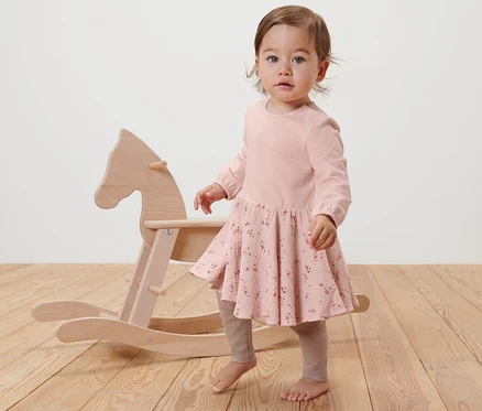 Un bébé se tient à côté d'un cheval à bascule en bois et porte une robe tissée rose.