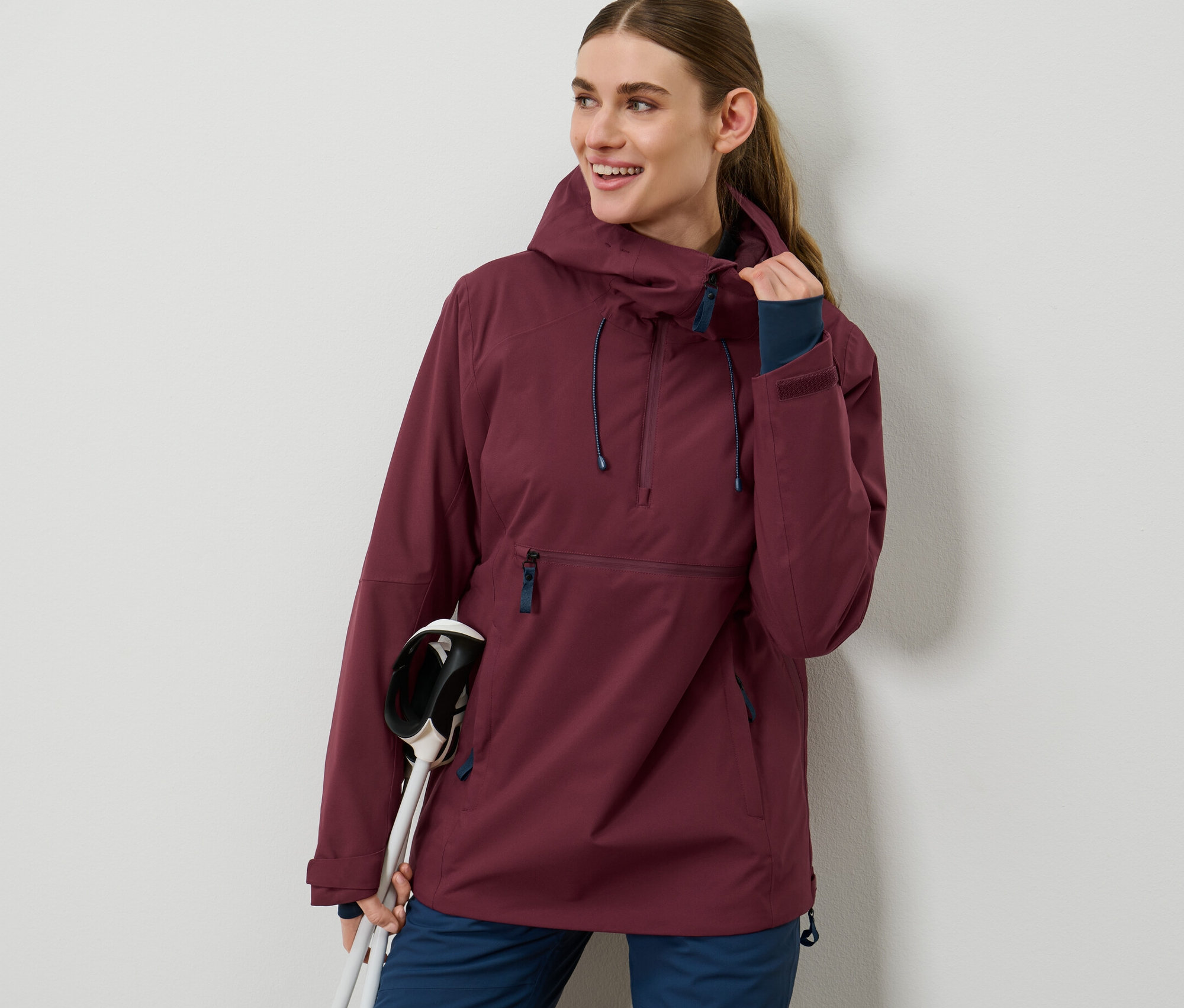 Une femme porte une veste de ski bordeaux à demi-fermeture éclair et tient des bâtons de ski.