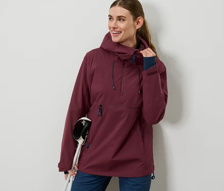 Une femme porte une veste de ski bordeaux à demi-fermeture éclair et tient des bâtons de ski.