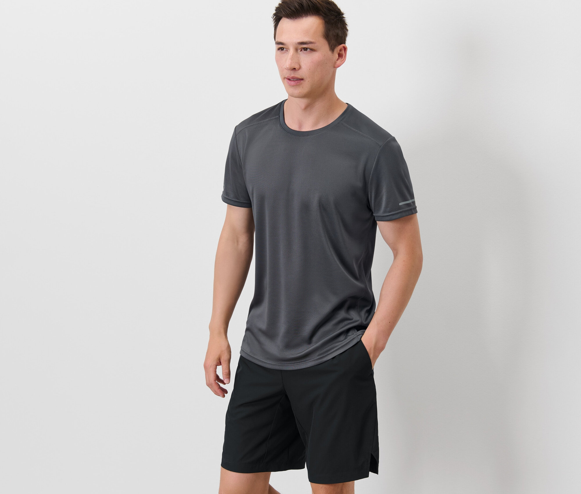Un homme se tient debout, les mains dans les poches de son short noir, portant un t-shirt technique gris.