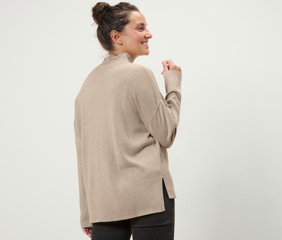 Femme regardant par-dessus son épaule portant un pull en maille fine beige avec col montant.