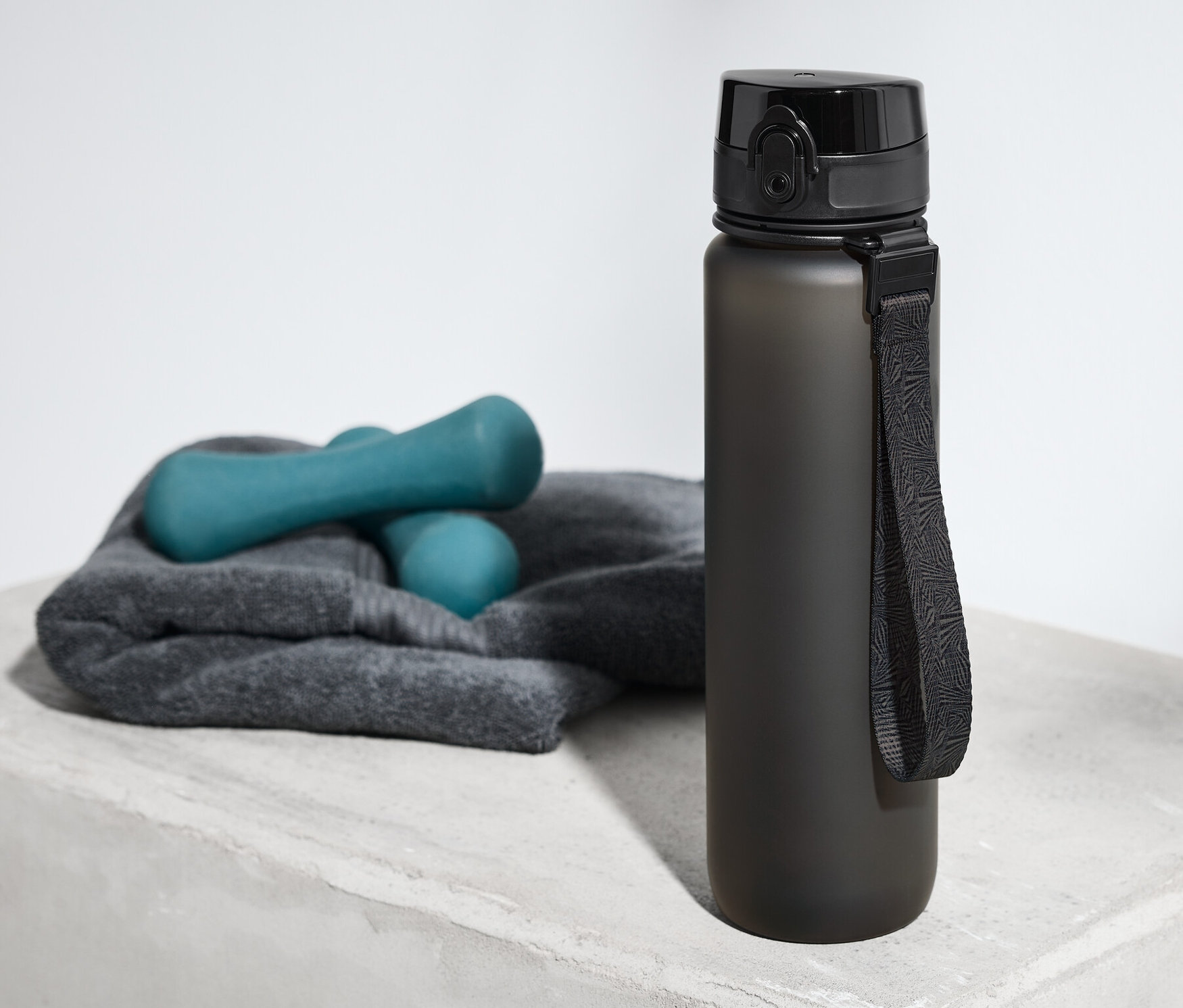 Une gourde noire est posée sur une surface grise à côté d'une serviette et d'haltères.