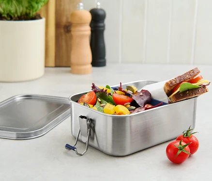Une boîte repas à emporter en acier inoxydable remplie de salade et d'un sandwich, avec deux tomates à côté.