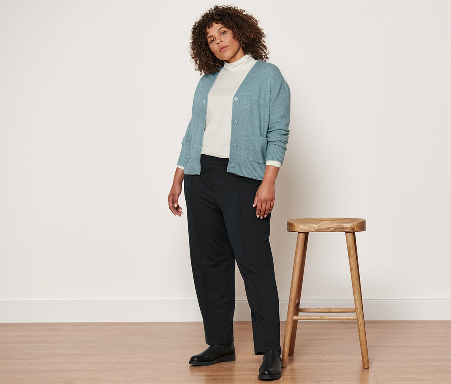 Une femme debout à côté d'un tabouret en bois porte une veste en maille avec de la laine, un t-shirt à col roulé crème, un pantalon en jersey noir avec nervure et des bottines en cuir.
