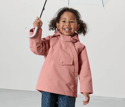 Une petite fille souriante tient un parapluie transparent au-dessus de sa tête. Elle porte une veste de pluie thermique rose pour tout-petit et un pantalon avec un motif de gouttes.