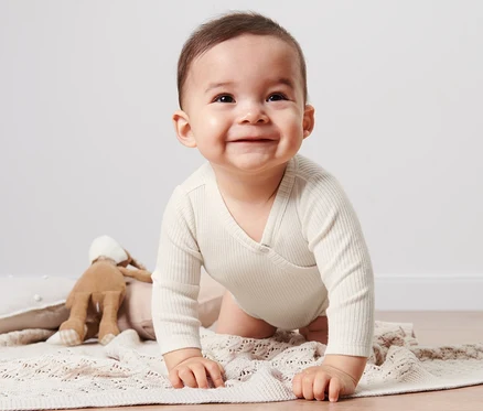 Bébé souriant en body avec modal pour bébé rampant sur une couverture tricotée beige à côté d'un lapin en peluche.
