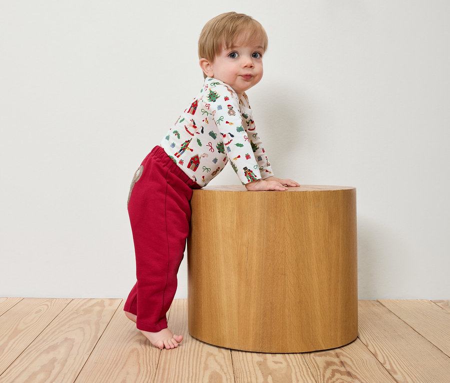 Un bébé est appuyé sur un tabouret en bois, portant un body de Noël et un pantalon rouge.