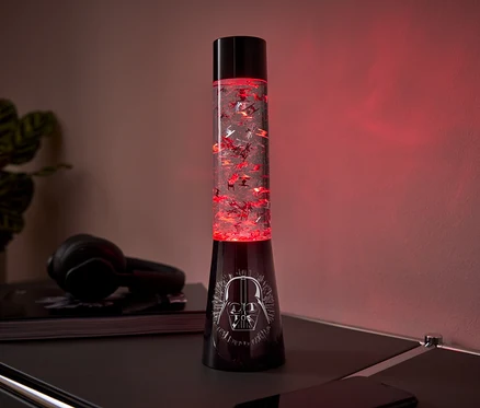 Une lampe à lave noire avec un motif Dark Vador sur la base et des paillettes rouges à l'intérieur, posée sur une table.