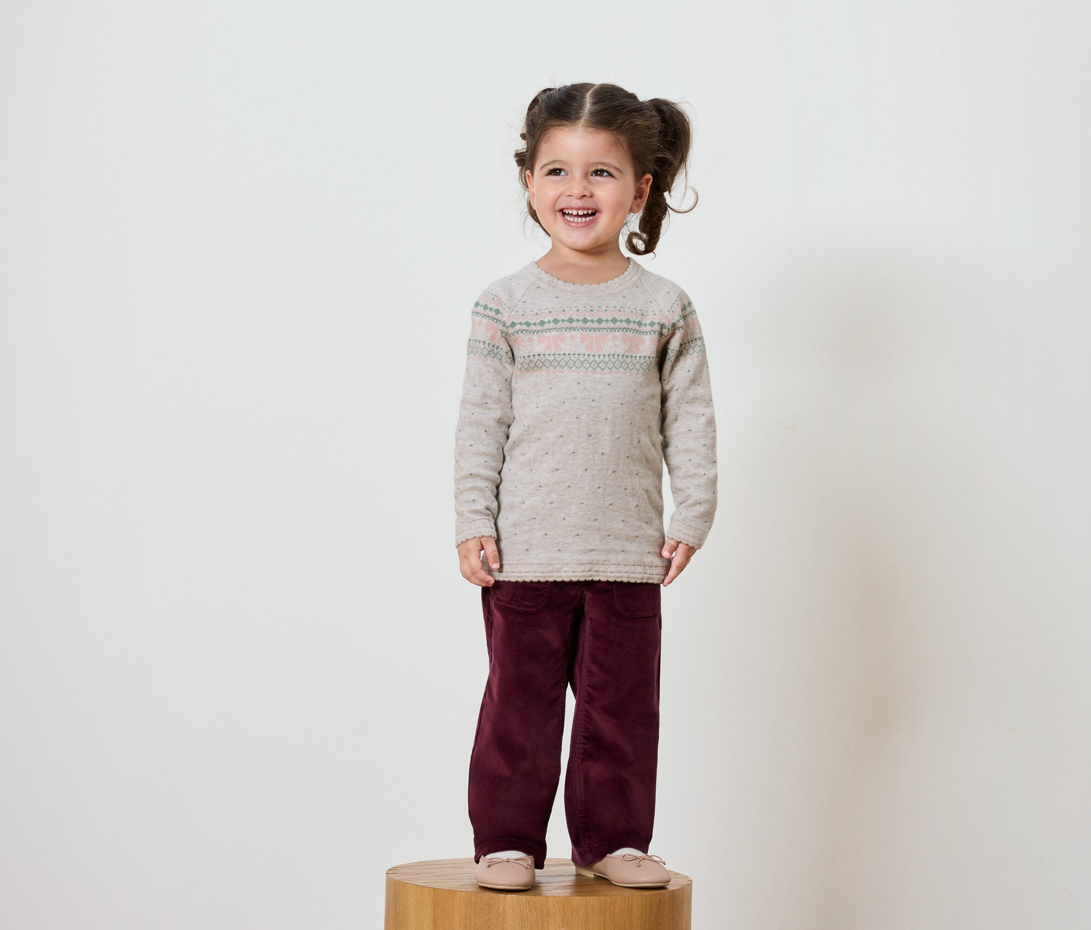 Petite fille souriante aux cheveux bruns avec des couettes debout sur un socle en bois. Elle porte un pull beige et un pantalon bordeaux.