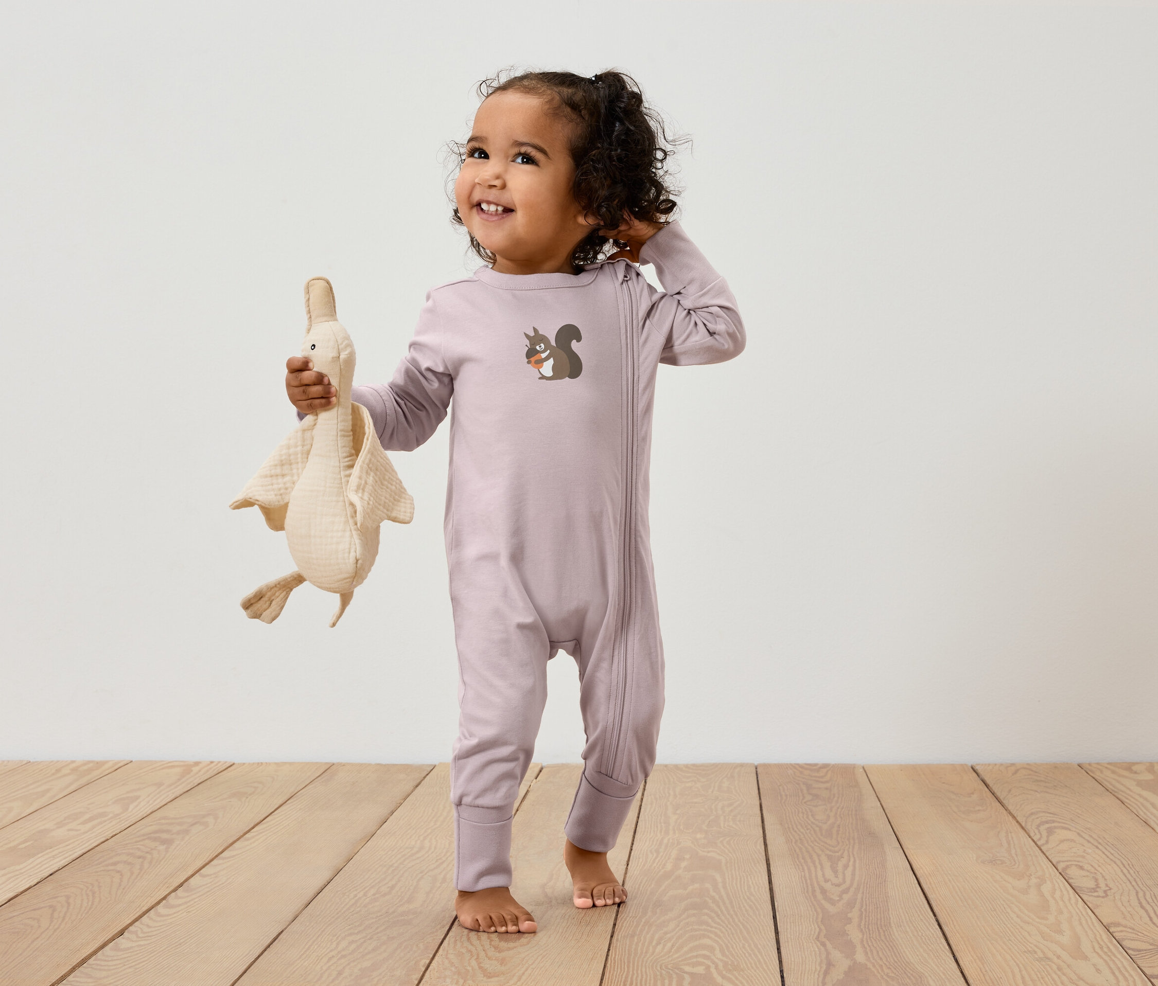 Un enfant dans une combinaison rose avec une fermeture éclair et une image d'écureuil tient une peluche d'oie.