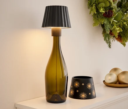 Sur une étagère blanche, une lampe faite d'une bouteille vert olive, un bougeoir noir avec des étoiles dorées, des décorations sur un plateau et une couronne de Noël en arrière-plan.