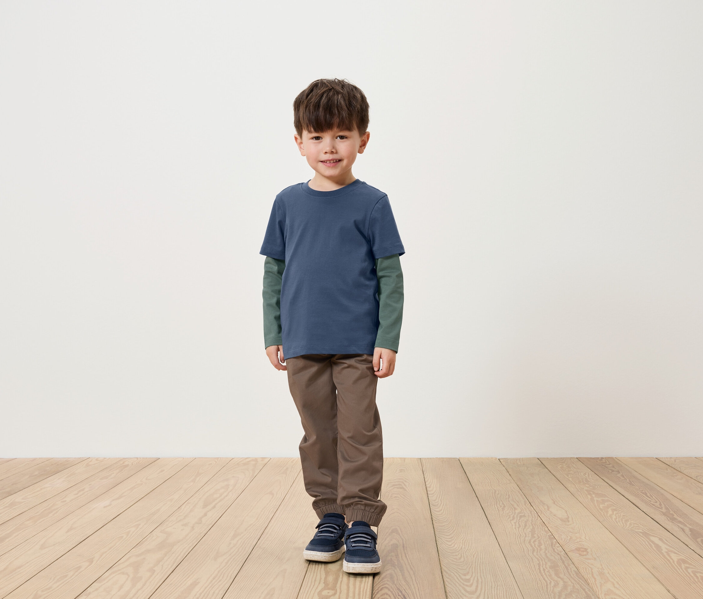 Un garçon se tient sur un plancher en bois, portant un t-shirt bleu à manches longues, un pantalon marron et des chaussures bleues.