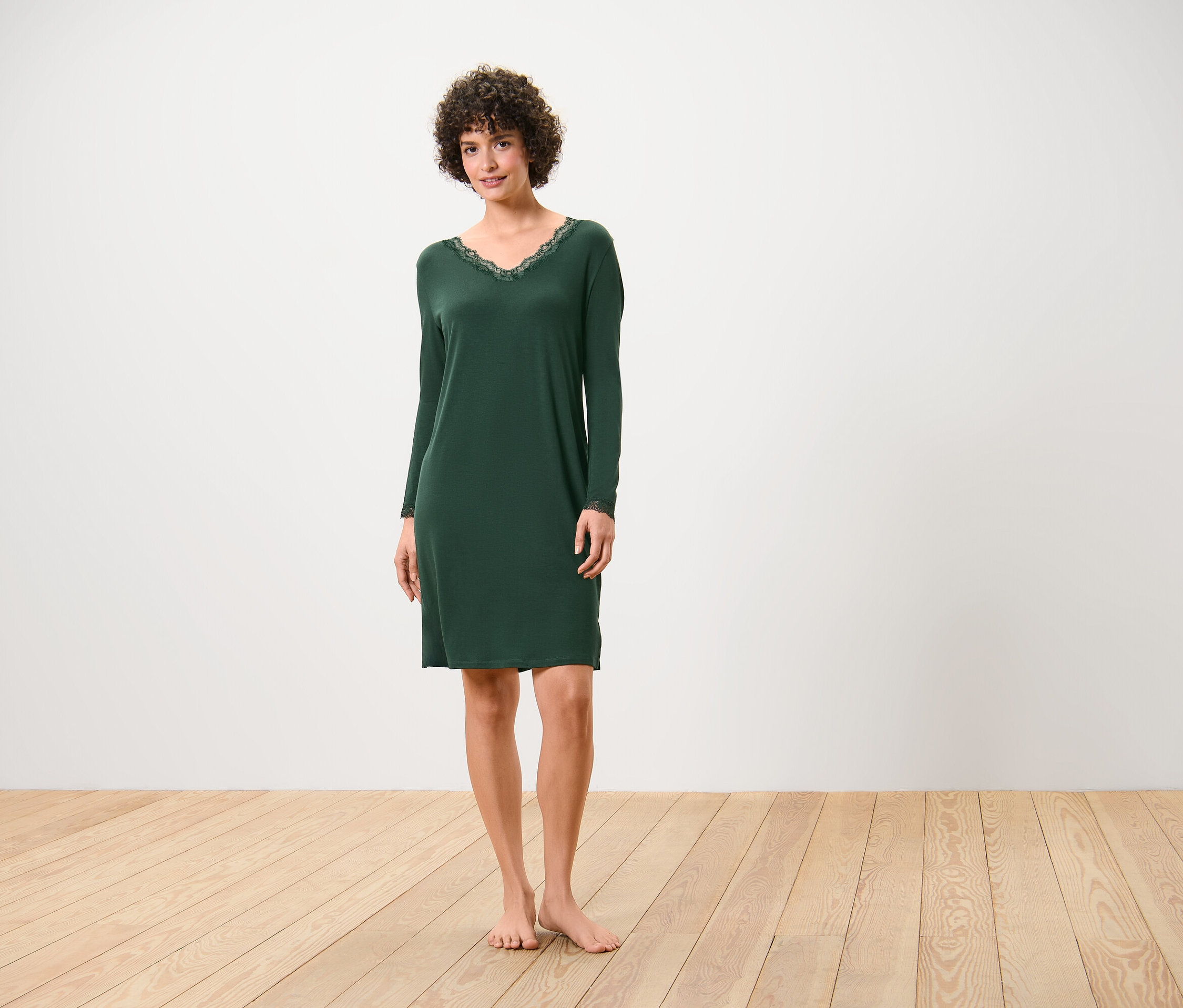 Une femme se tient pieds nus sur un plancher en bois, portant une chemise de nuit vert foncé à manches longues avec de la dentelle à l'encolure.