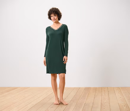 Une femme se tient pieds nus sur un plancher en bois, portant une chemise de nuit vert foncé à manches longues avec de la dentelle à l'encolure.