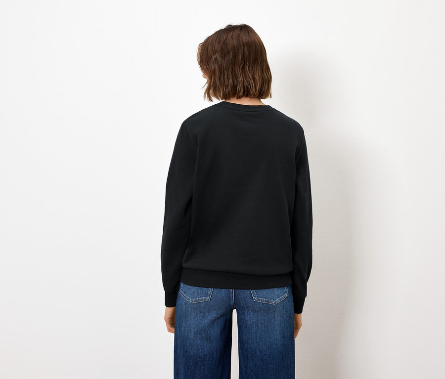 De dos, une femme aux cheveux bruns porte un jean bleu et un pull noir.