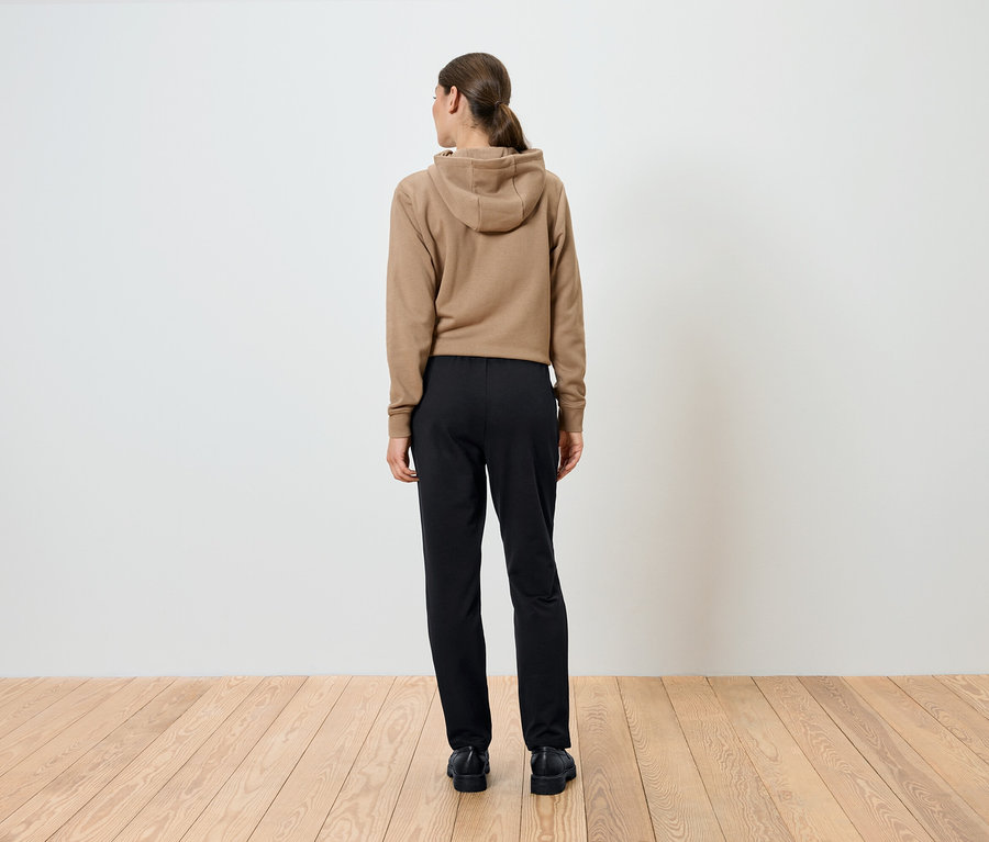 Femme portant un sweat à capuche marron et un pantalon noir, dos à la caméra.