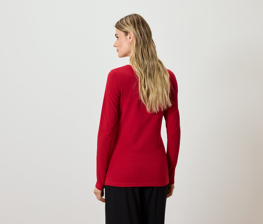 Une femme blonde se tient dos à la caméra, portant un chemisier rouge à manches longues et un pantalon noir.