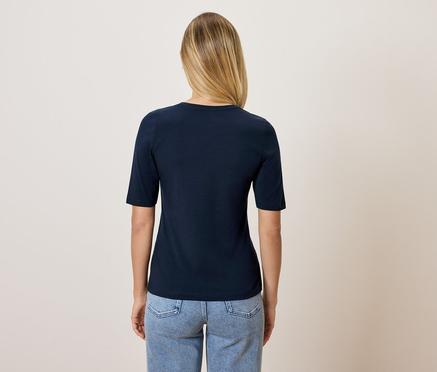 Une femme aux cheveux blonds, vue de dos, porte un t-shirt bleu marine et un jean bleu.