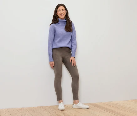 Une femme debout portant un col roulé violet, un legging marron et des baskets blanches.