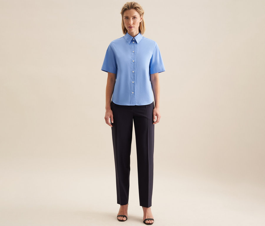 Une femme debout de face porte un chemisier à manches courtes Seidensticker bleu clair, un pantalon foncé et des sandales noires.
