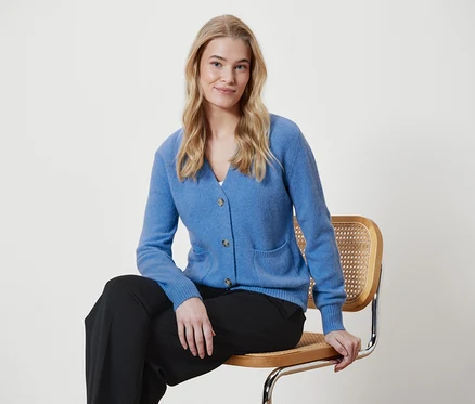 Une femme blonde est assise sur une chaise, portant un cardigan bleu et un pantalon noir.