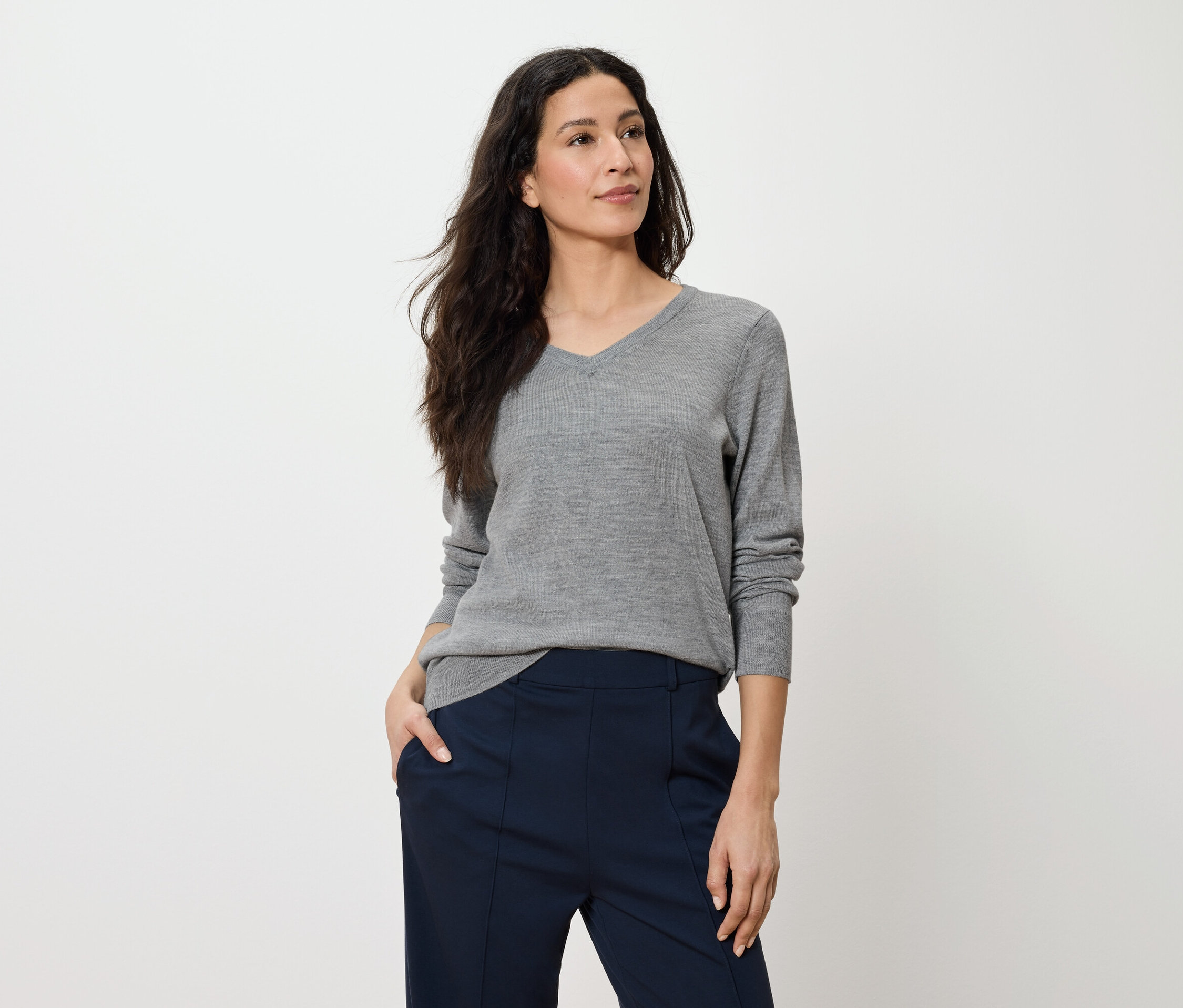 Une femme pose, portant un pull gris à manches longues et un pantalon bleu marine.