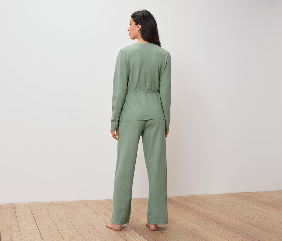Une femme en pull et pantalon verts se tient dos au spectateur sur un plancher en bois.