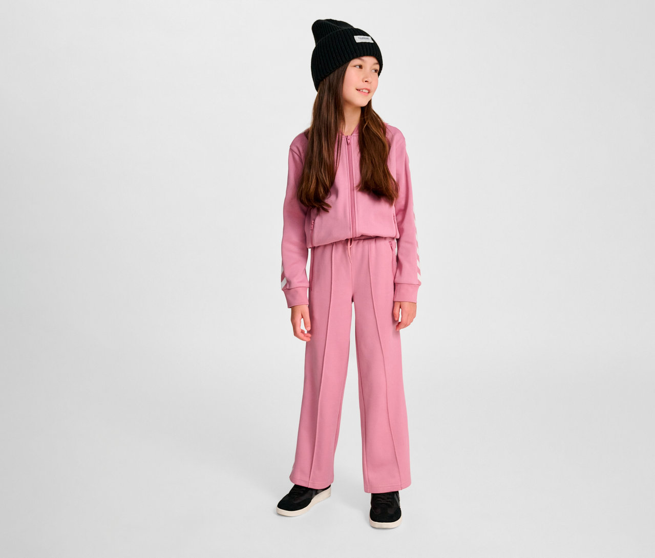Une fille portant un survêtement rose et un bonnet noir. Elle porte un pantalon HUMMEL HMLJR Archive Wideleg Poly.
