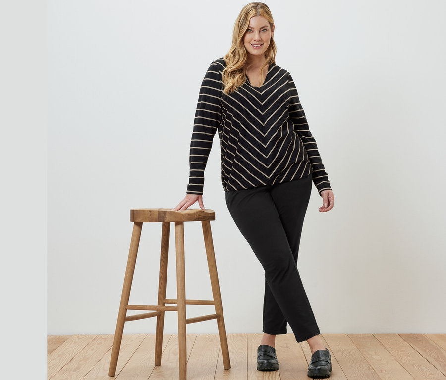 Une femme pose, appuyée sur un tabouret en bois, portant une blouse rayée en modal mélangé et un pantalon de jogging noir.