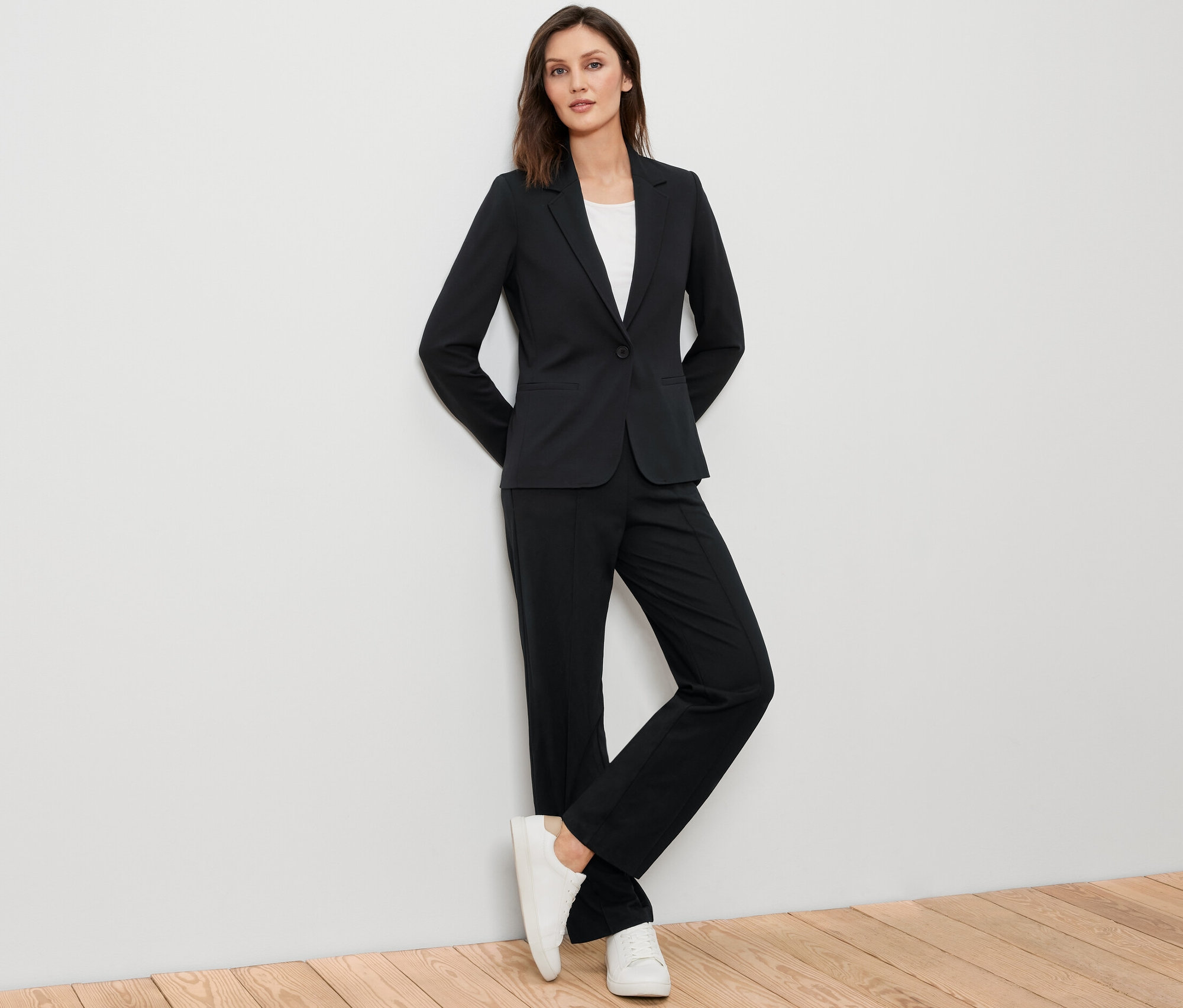 Femme posant en blazer en jersey noir, pantalon noir et baskets blanches.