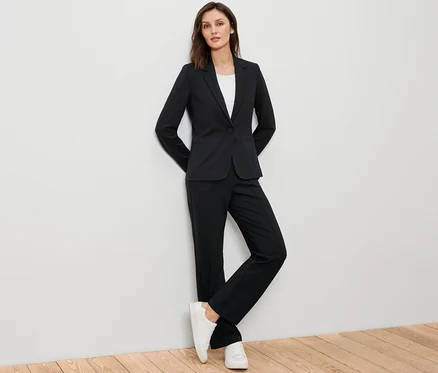 Femme posant en blazer en jersey noir, pantalon noir et baskets blanches.