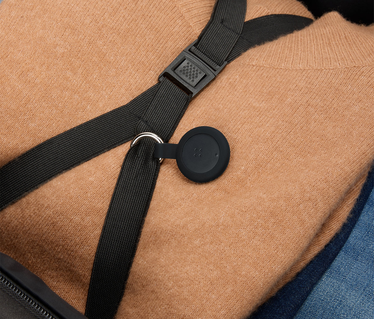 Un pull marron et un jean bleu sont placés dans une valise, avec une sangle noire et un pendentif rond noir par-dessus.