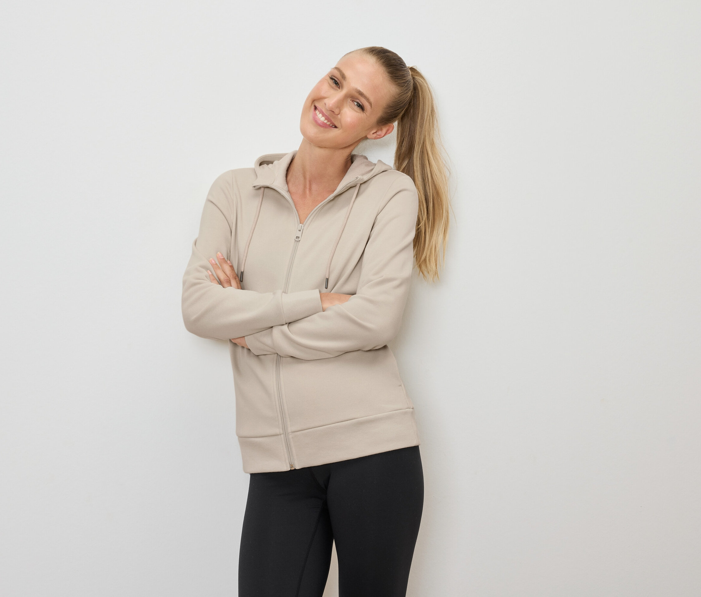 Une femme blonde souriante avec une queue de cheval pose les bras croisés, portant un sweat à capuche beige zippé et un legging noir.