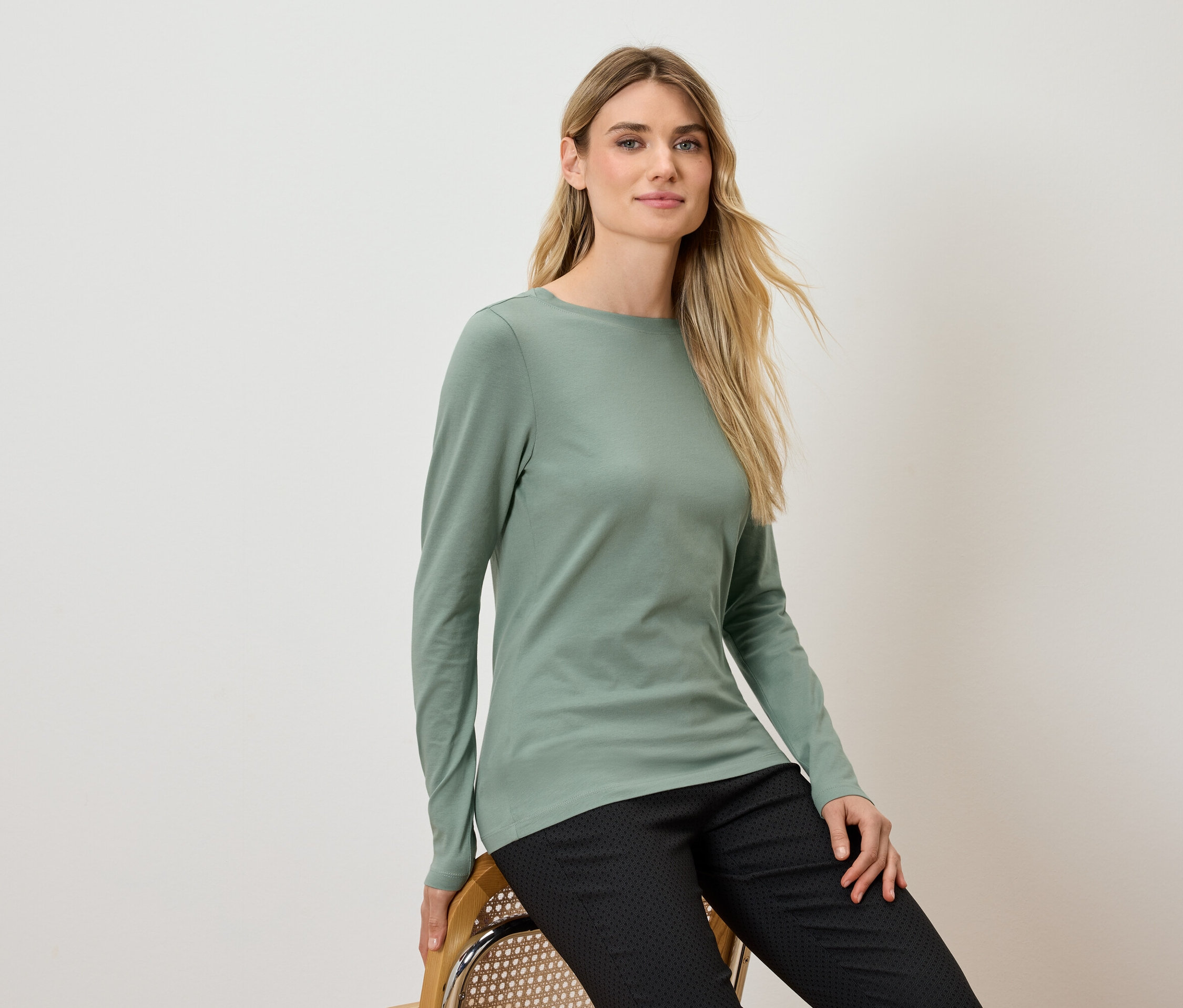 Une femme blonde est assise sur une chaise, portant un T-shirt vert à manches longues et un pantalon noir.