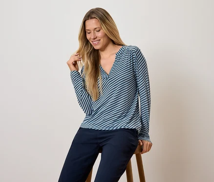 Une femme est assise sur un tabouret, portant un haut à manches longues rayé bleu et blanc et un pantalon bleu foncé.