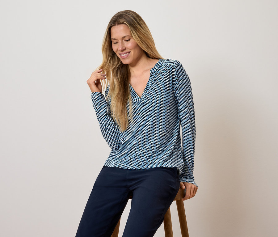 Une femme est assise sur un tabouret, portant un haut à manches longues rayé bleu et blanc et un pantalon bleu foncé.