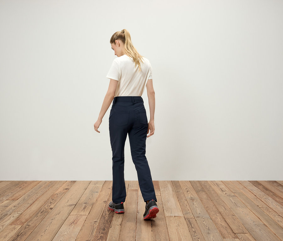 De dos, on voit une femme blonde avec une queue de cheval, un t-shirt blanc, un pantalon bleu marine et des baskets.