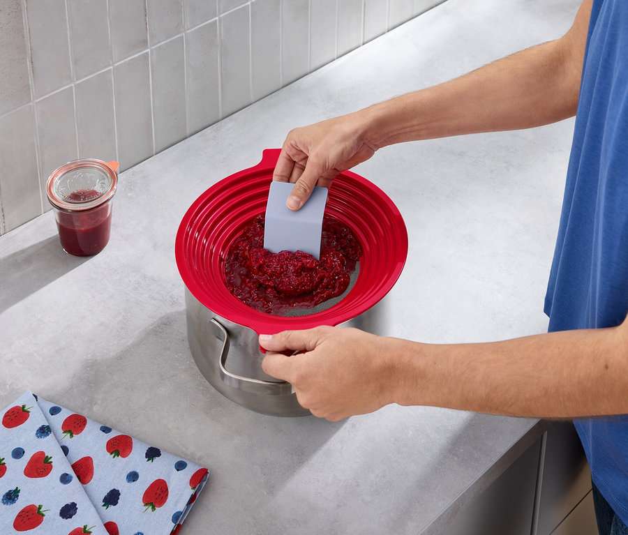 Un homme passe de la confiture de framboises dans une passoire pour farine et pour tamiser au-dessus d'une casserole avec une spatule grise.