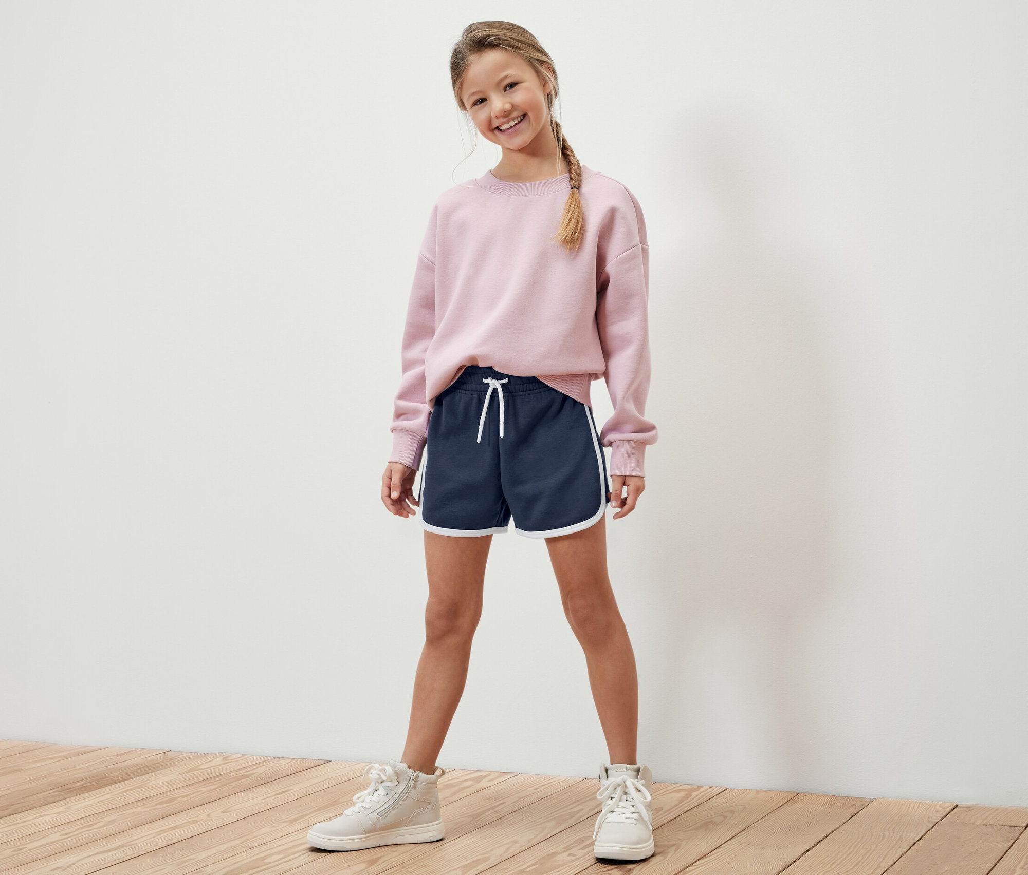 Une fille pose en pull rose et short de survêtement bleu marine avec bordure blanche. Elle porte 2 shorts de survêtement pour enfant.
