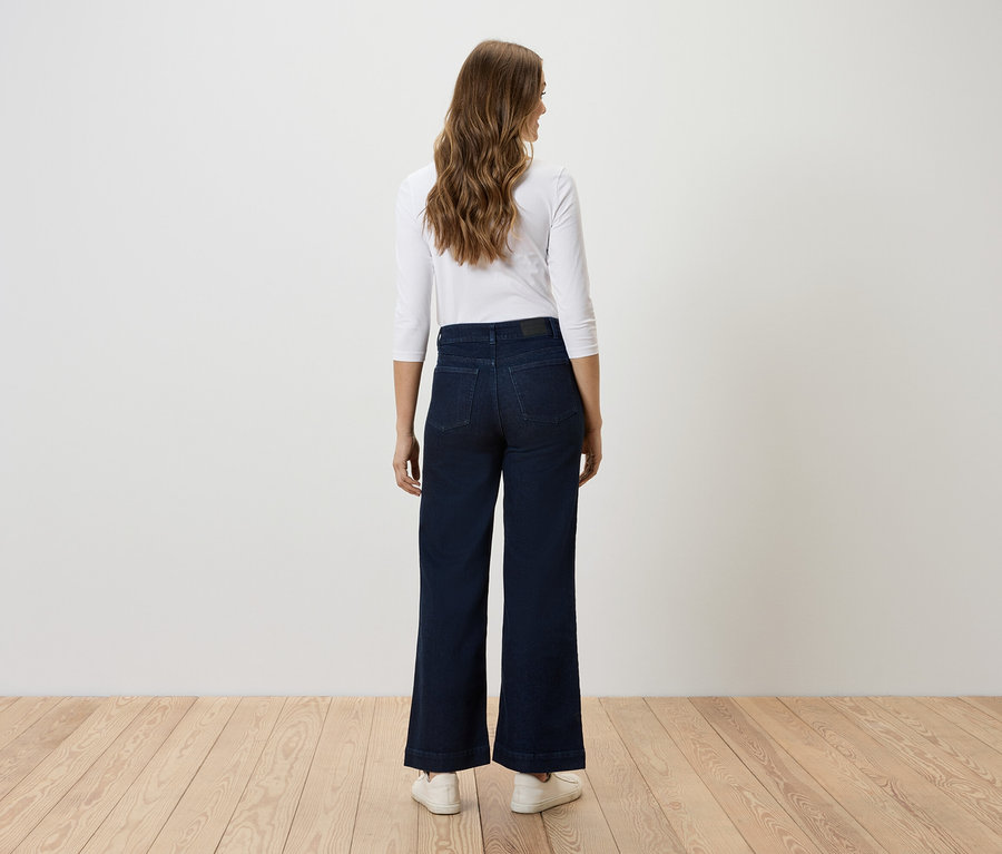 Une femme se tient dos à la caméra, portant un jean large bleu foncé et un haut blanc.