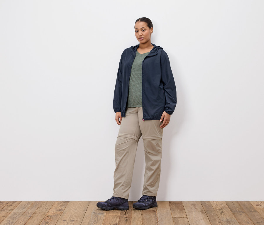Une femme se tient devant un mur blanc, portant une veste bleu marine, un t-shirt vert olive, un pantalon kaki et des chaussures bleu marine.