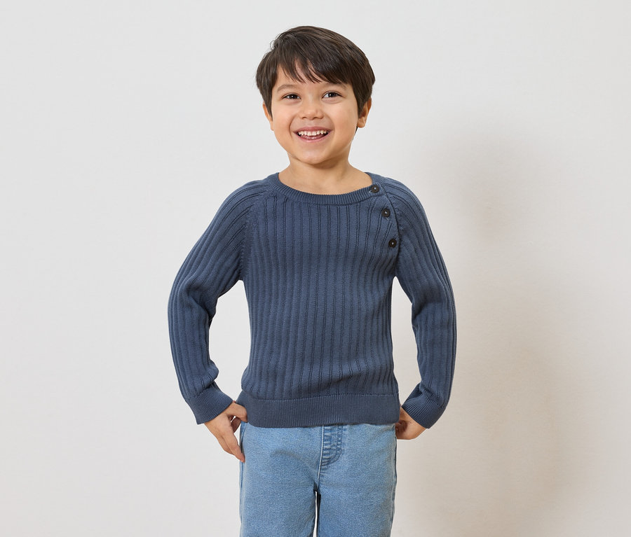 Un garçon souriant aux cheveux bruns porte un pull bleu marine côtelé avec des boutons et un jean bleu clair.