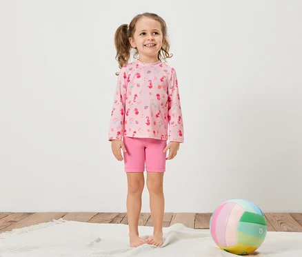Une fillette avec des tresses porte un t-shirt rose à manches longues avec un motif d'hippocampes et un short rose. Un ballon coloré repose sur un tapis blanc.