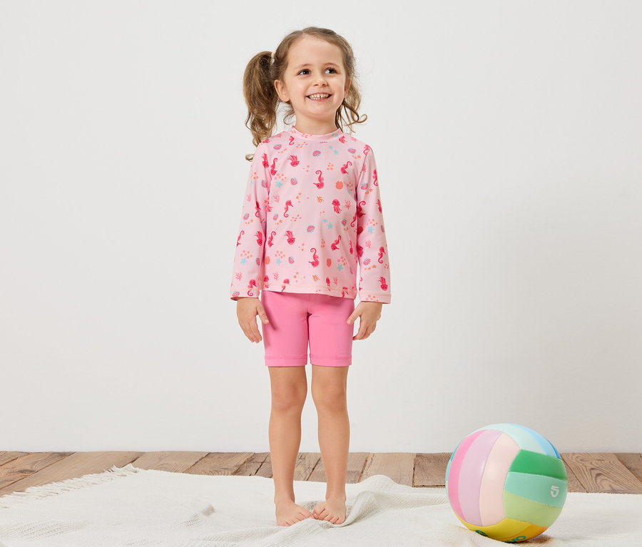Une fillette avec des tresses porte un t-shirt rose à manches longues avec un motif d'hippocampes et un short rose. Un ballon coloré repose sur un tapis blanc.