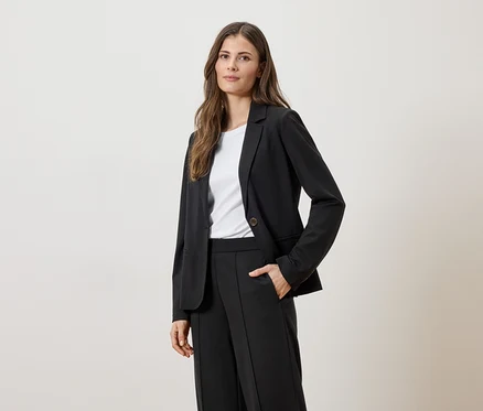Une femme aux cheveux bruns porte un veston noir sur un t-shirt blanc et un pantalon noir, une main dans la poche.