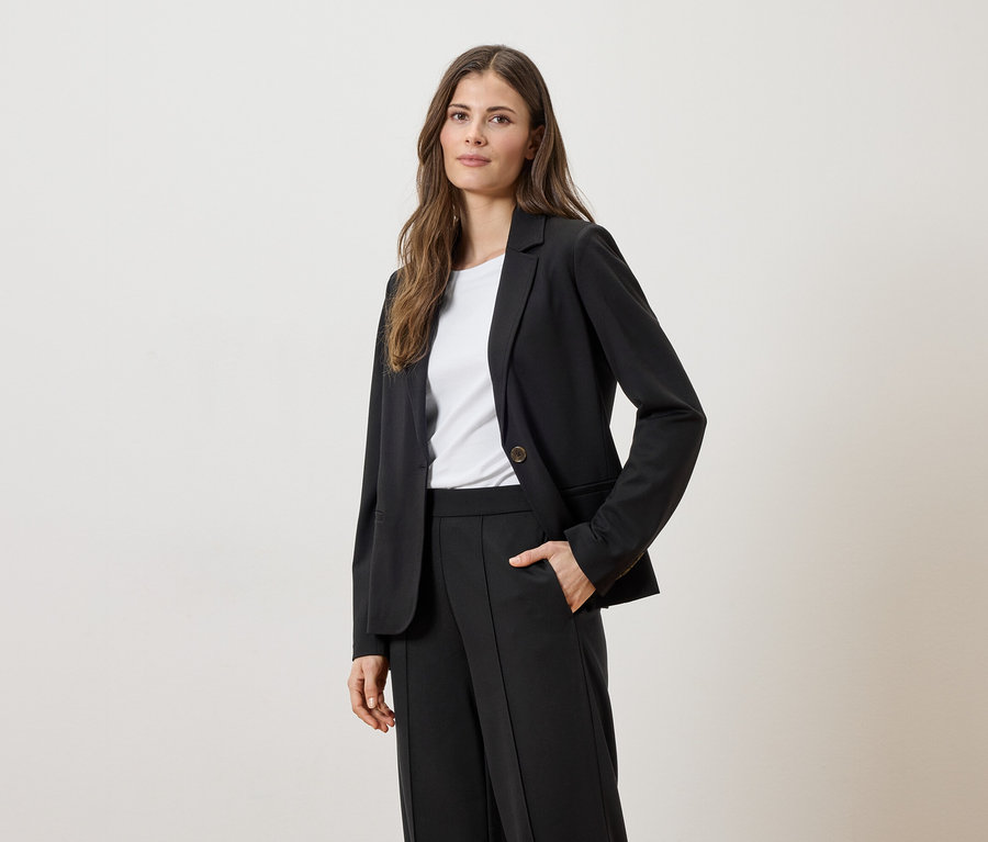 Une femme aux cheveux bruns porte un veston noir sur un t-shirt blanc et un pantalon noir, une main dans la poche.