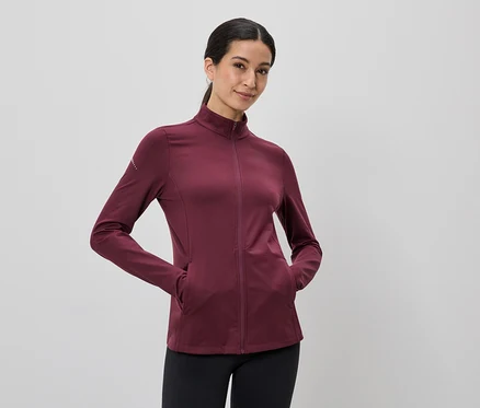 Une femme portant une queue de cheval porte une veste bordeaux zippée et un legging noir, les mains dans les poches.