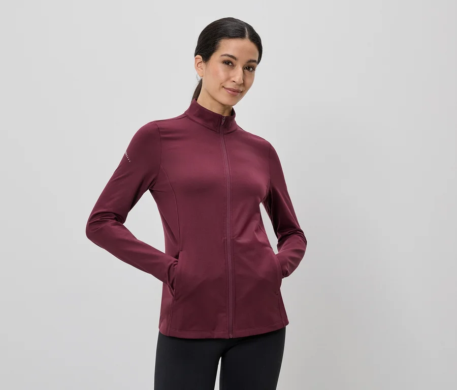 Une femme portant une queue de cheval porte une veste bordeaux zippée et un legging noir, les mains dans les poches.