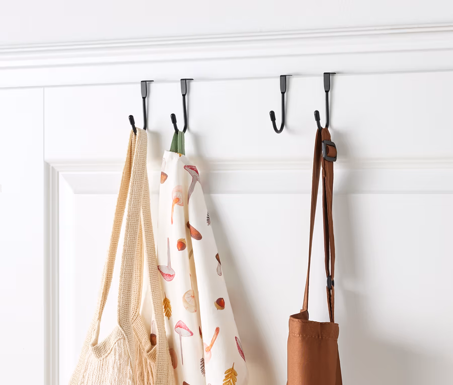 Sur une porte blanche, quatre crochets noirs soutiennent un sac en filet rempli de fruits, un torchon à motifs de champignons et un tablier marron.